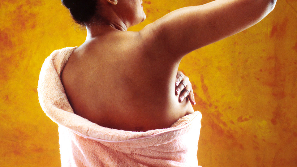 A woman seen from behind, with a towel around her body, holding her hand over one breast.